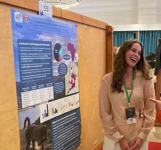White woman with long brown hair smiling and talking to another person out of frame while presenting scientific poster