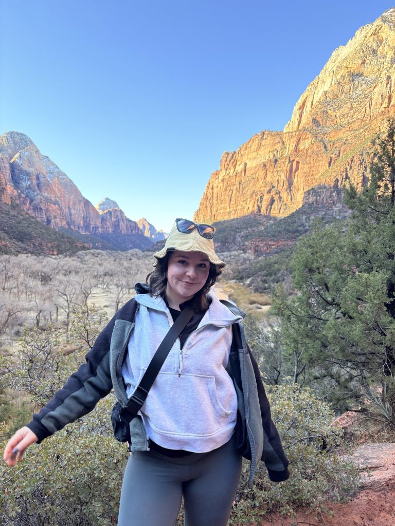 White woman with brown hair wearing hat standing in front of a mountain